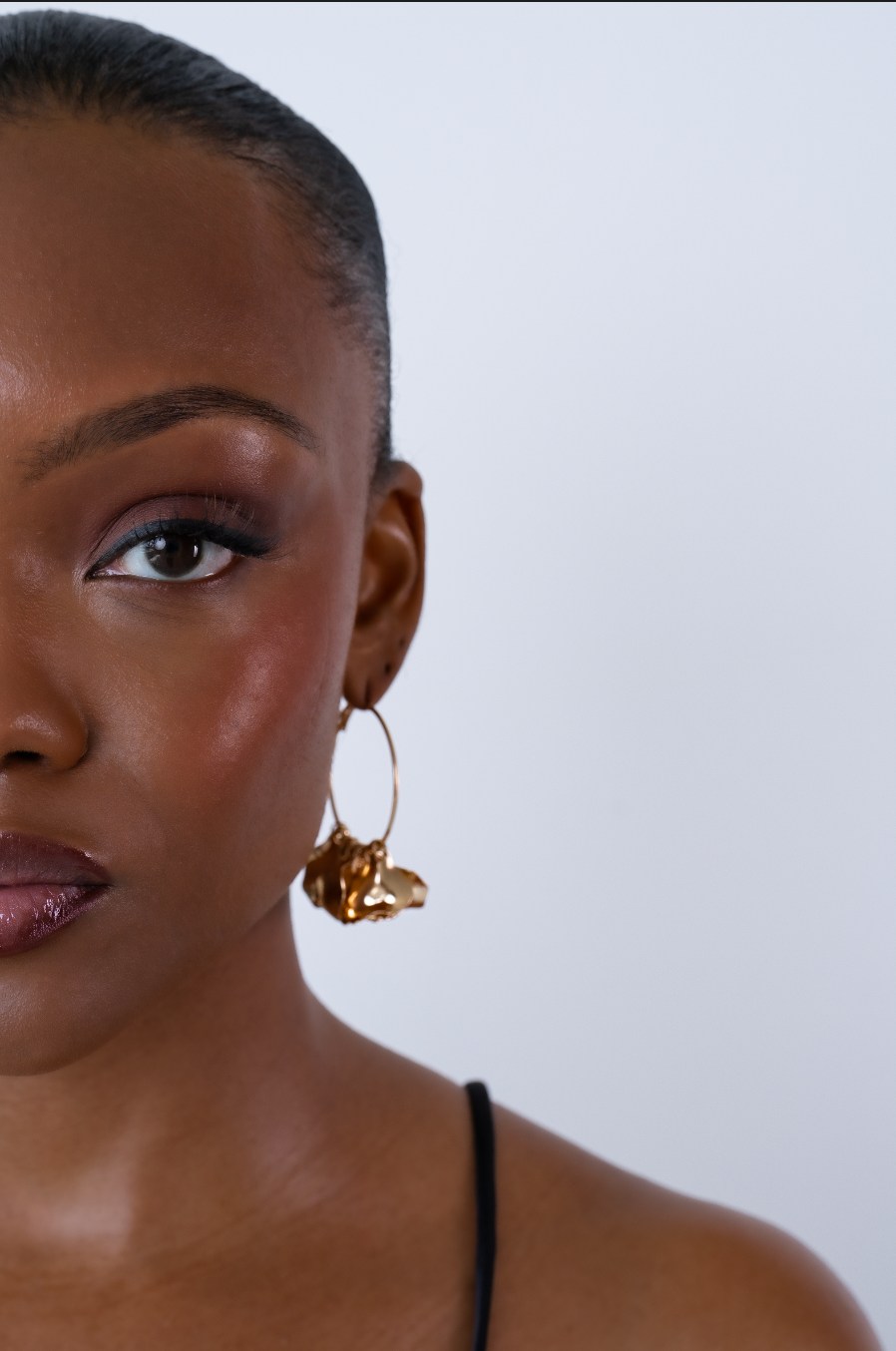 Close-up of a woman wearing a gold hoop earring with an abstract charm, featuring bold eye makeup and short hair.