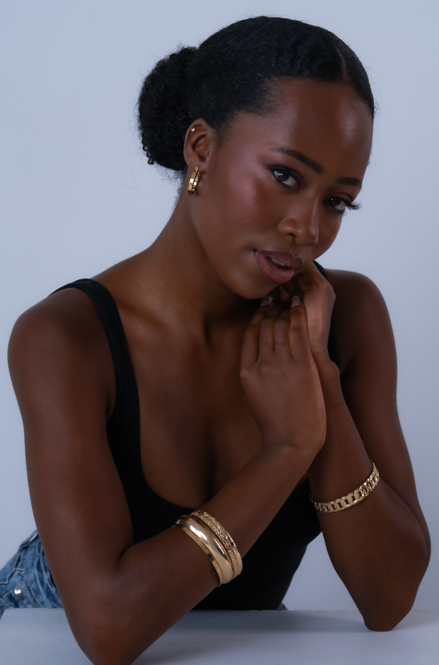 Woman in black tank top posing with gold bracelets and hoop earrings against a light gray background.