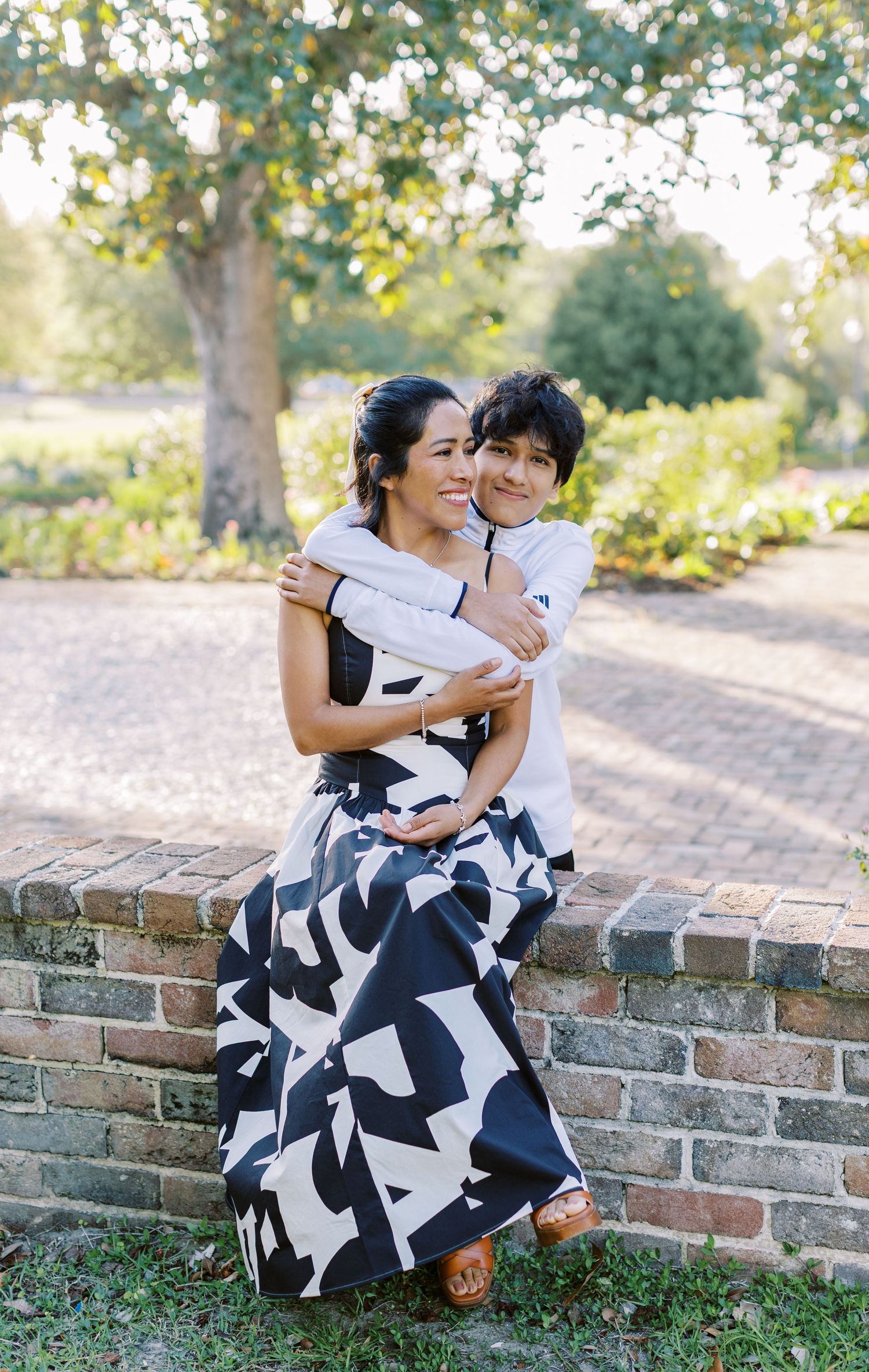A woman in a black and white printed maxi dress sits on a brick wall hugging a young boy in a park.