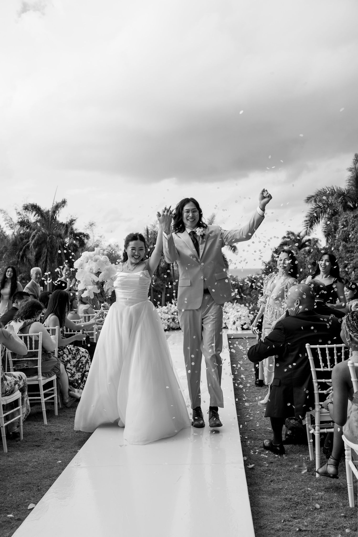 Black and white photo of joyful newlyweds walking down the aisle, arms raised, confetti flying at outdoor wedding.