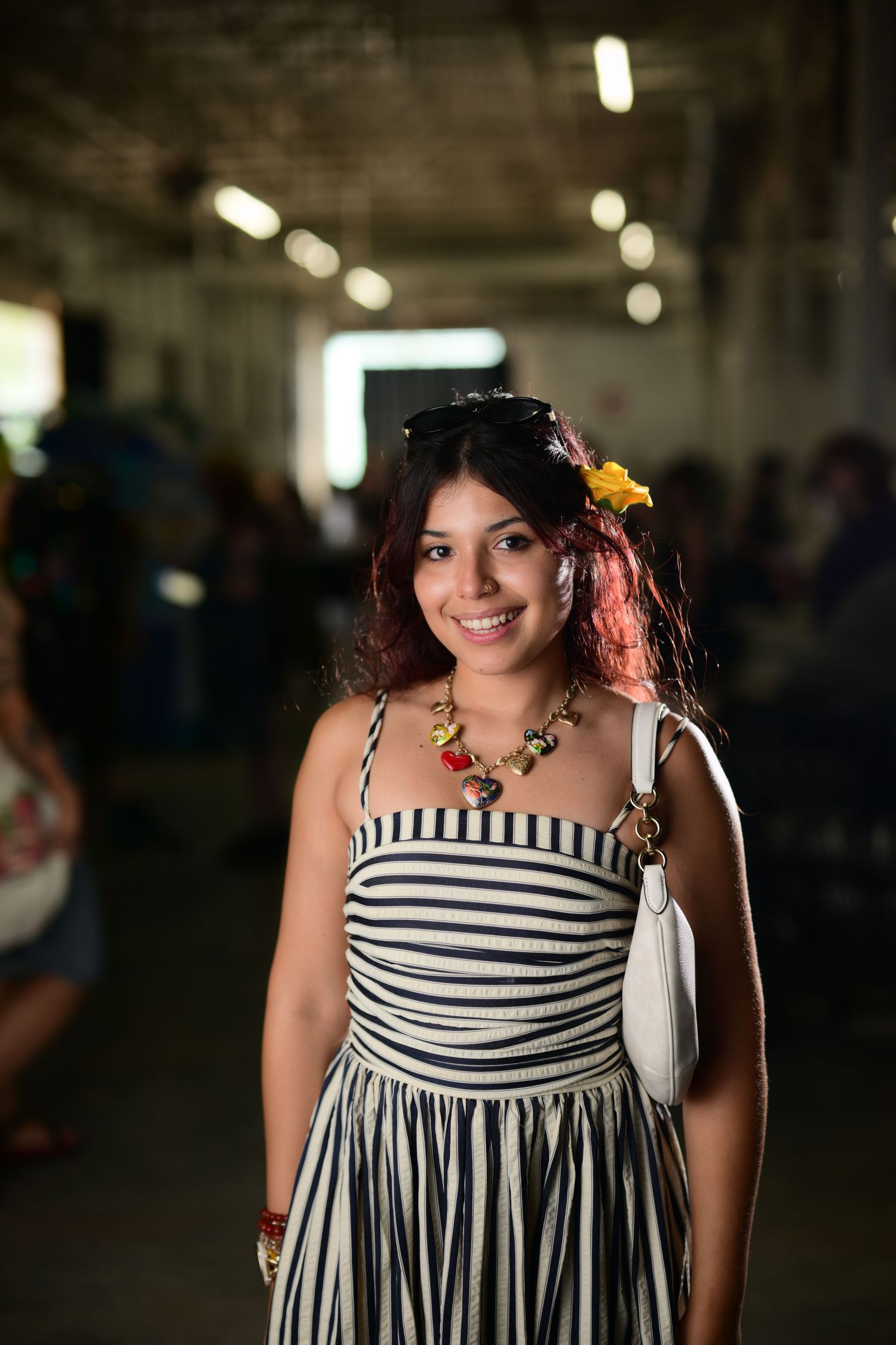 Smiling young woman in striped sundress with yellow flower in hair and white purse at indoor event.