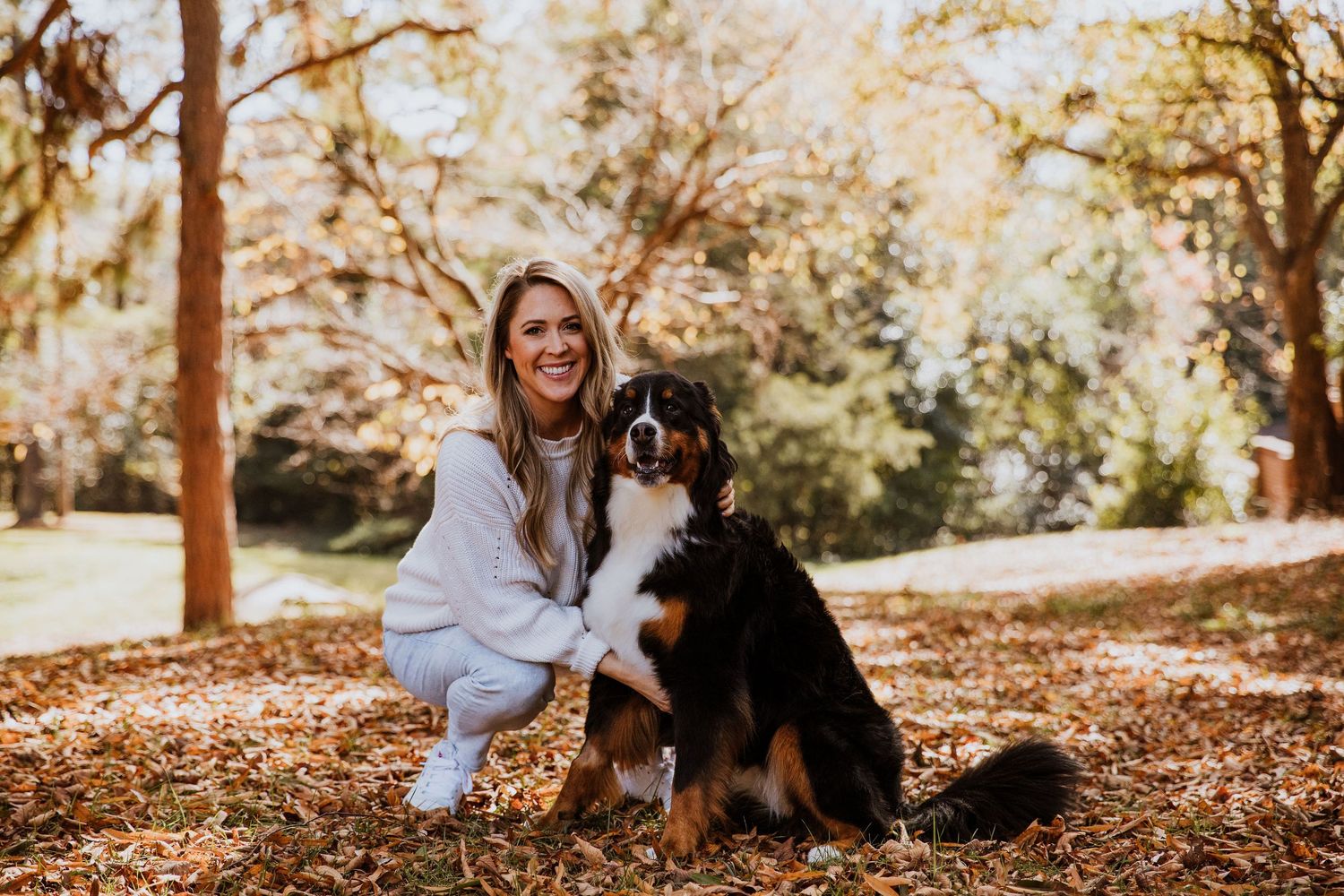 Woman smiling with a Bernese Mountain Dog in an autumn park surrounded by fallen leaves and trees.