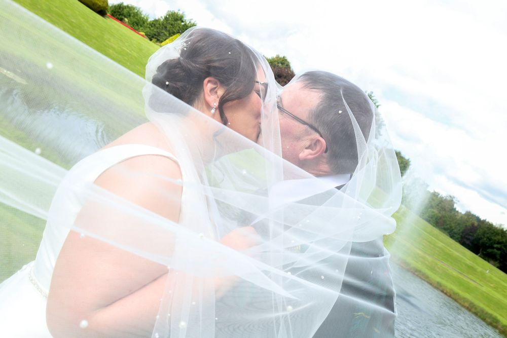 Bride and groom sharing a romantic kiss under a flowing white veil outdoors near a pond on their wedding day.