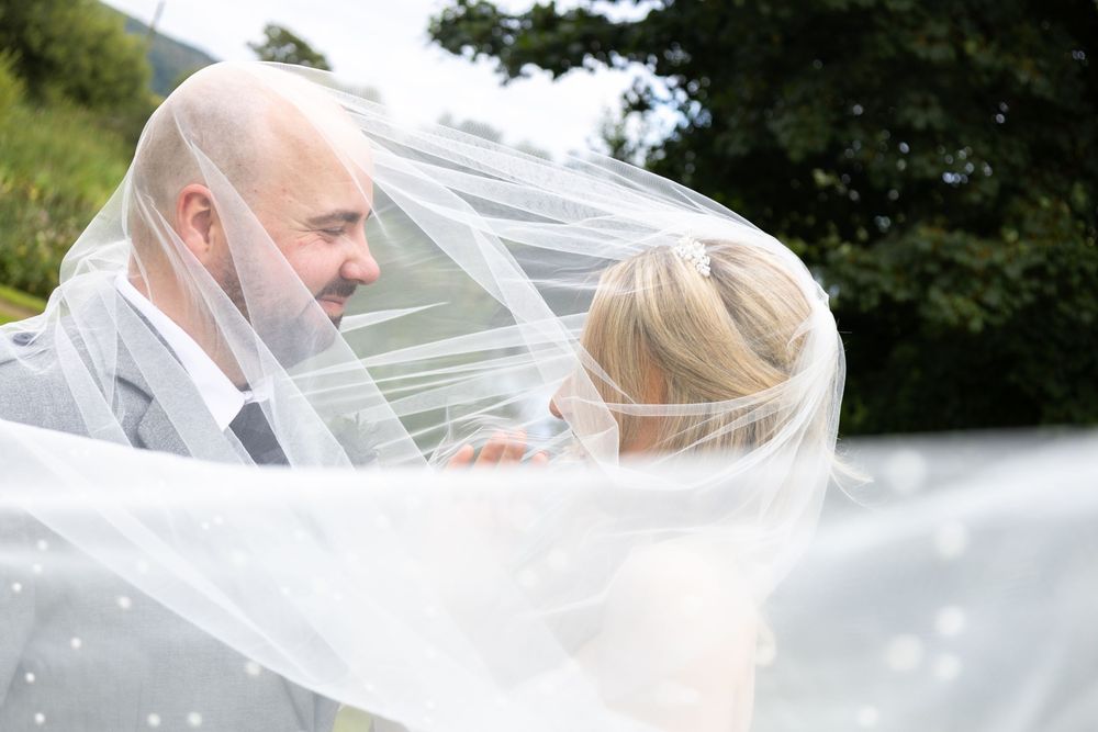 Bride and groom share romantic moment under flowing wedding veil outdoors.
