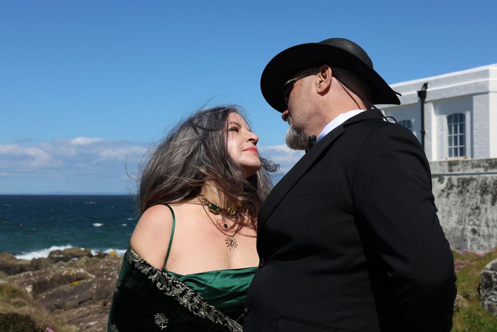 Couple in elegant attire sharing a romantic moment by the ocean, woman in green velvet dress, man in black suit and hat.