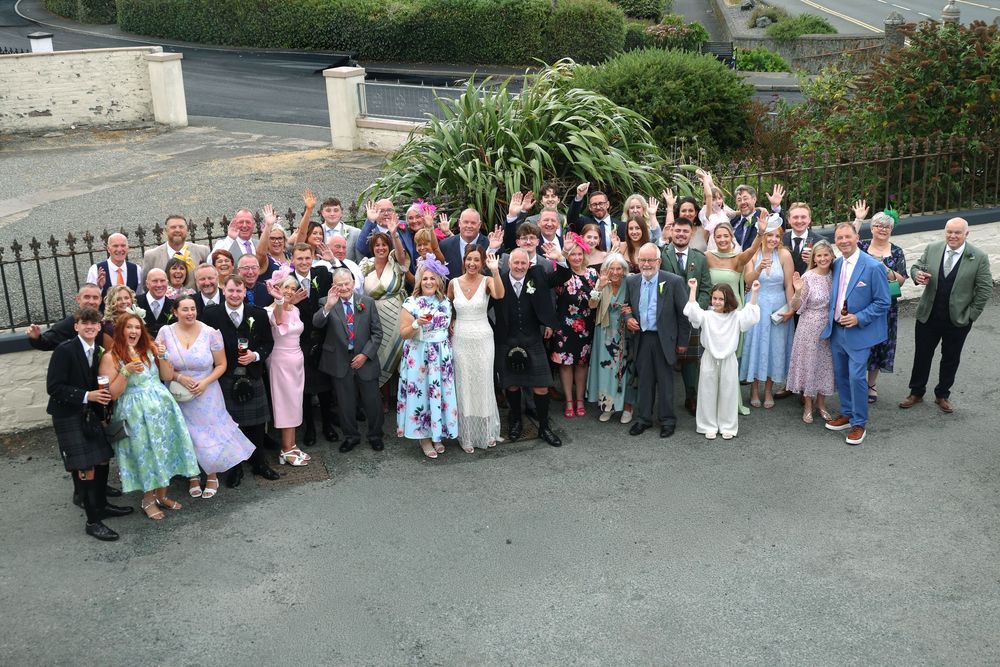 Large wedding group photo outdoors with bride and groom surrounded by guests in formal attire on a sunny day.
