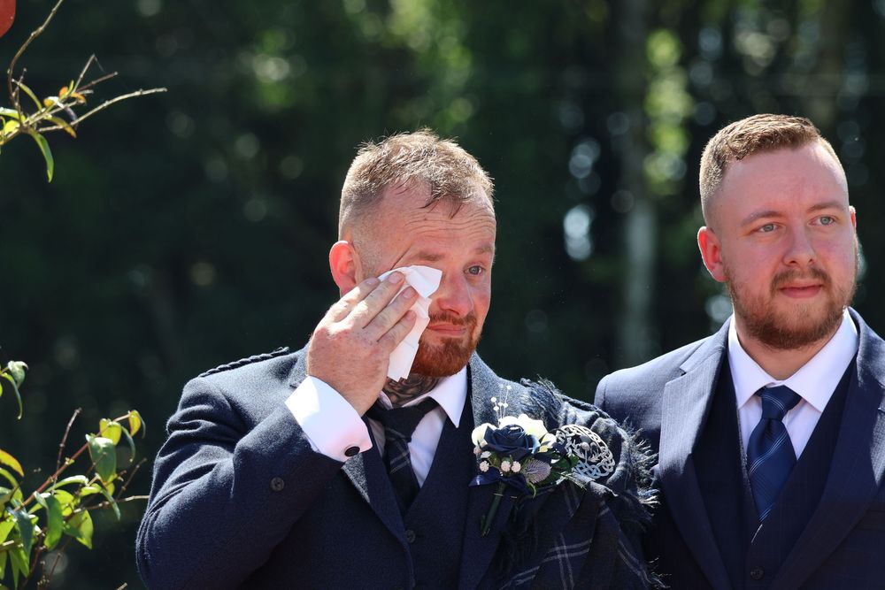 Emotional groom wiping tears with handkerchief at outdoor wedding ceremony, standing beside best man in kilt.