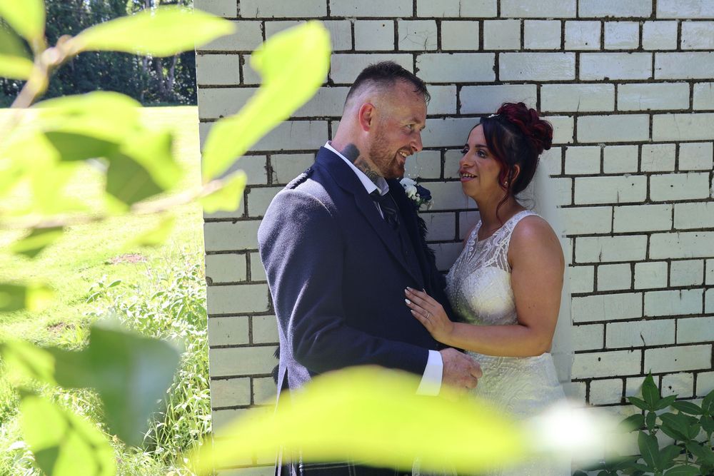 A bride and groom share a romantic moment against a brick wall, surrounded by lush greenery on their wedding day.