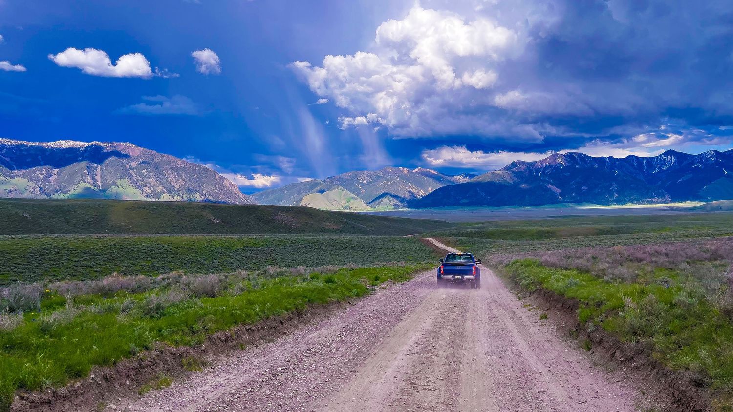 Blue truck driving on dirt road through vast green mountain valley.