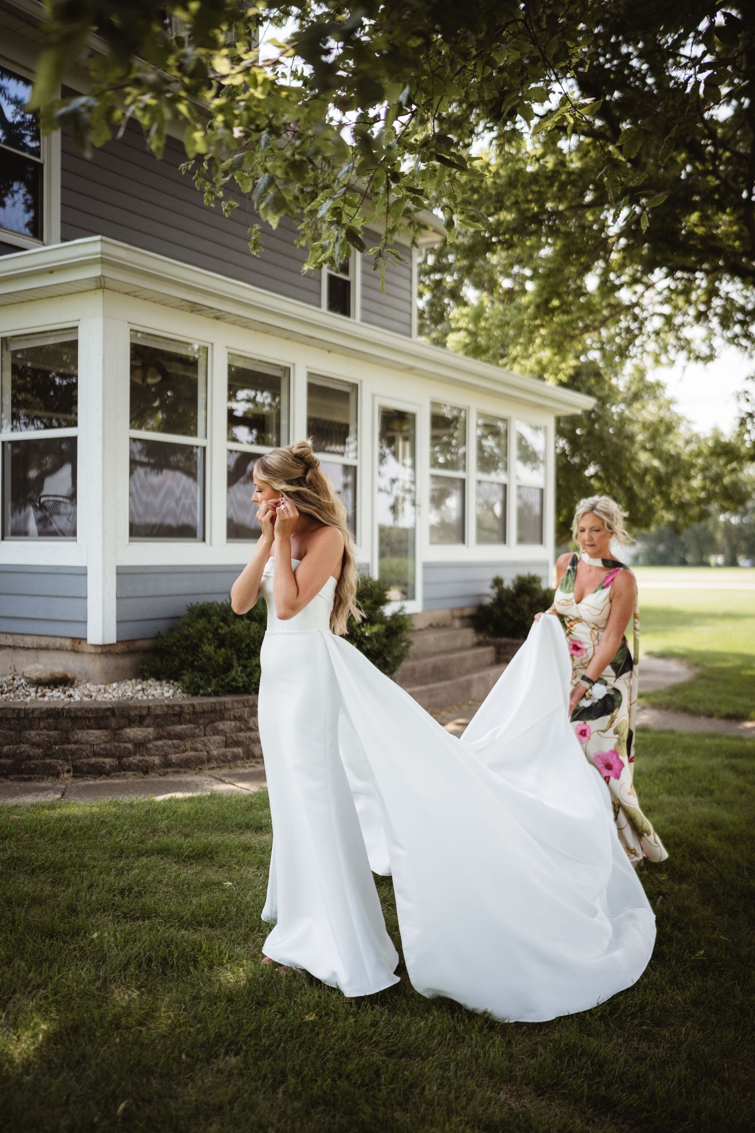 Bride in white gown being helped with train by bridesmaid outside gray house on green lawn.