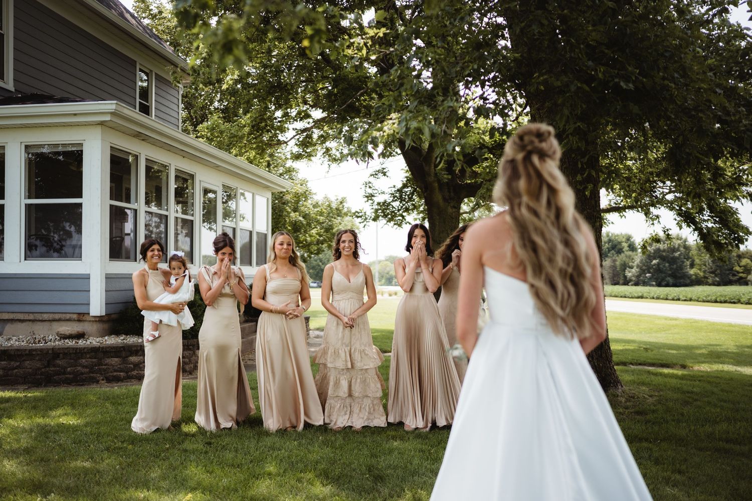 Bride in white gown reveals herself to bridesmaids in champagne dresses outdoors near a tree.