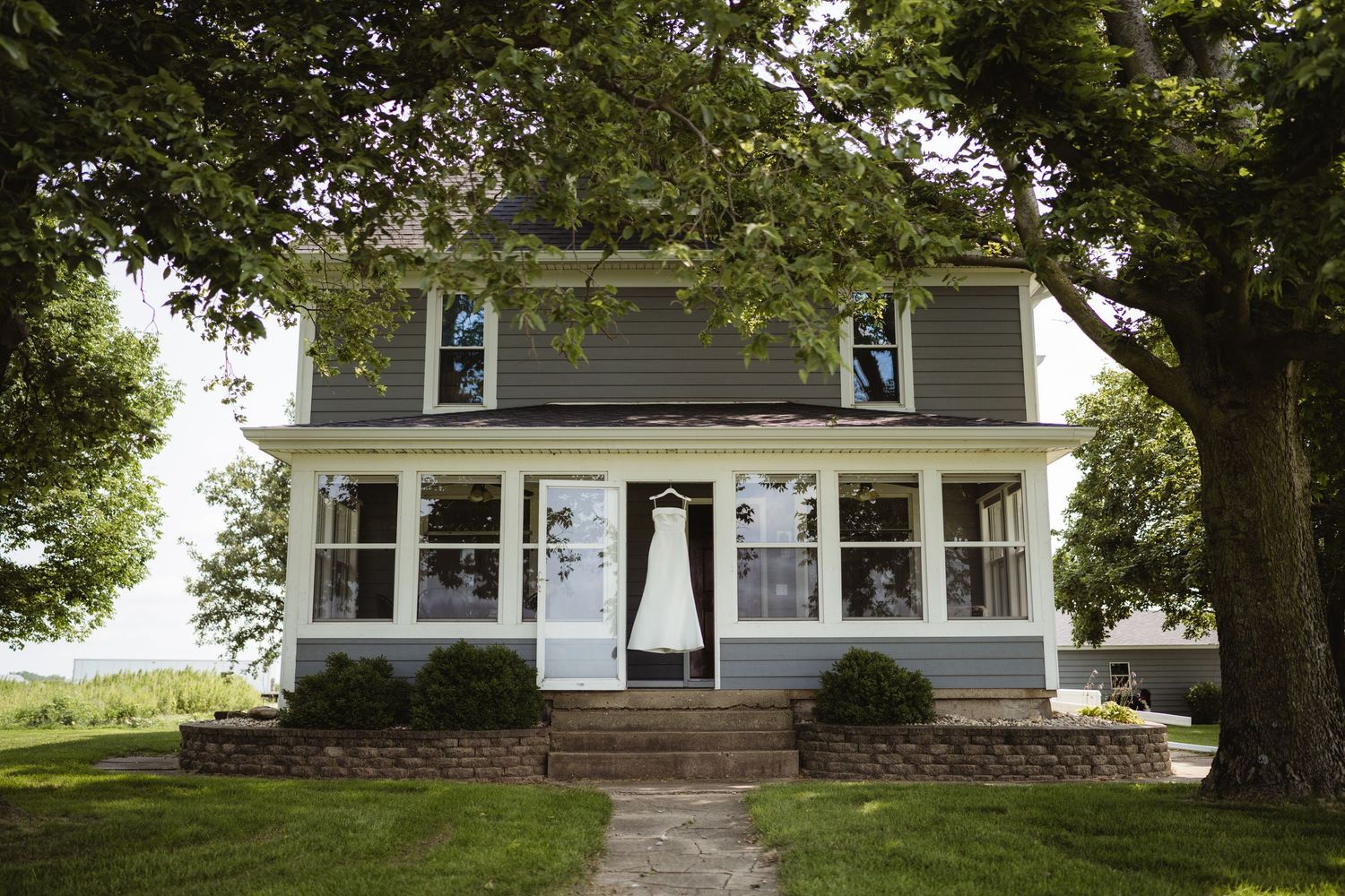 A white wedding dress hangs on a farmhouse porch surrounded by large trees and lush green lawn.