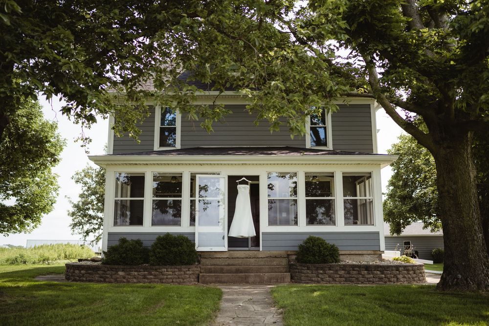 A white wedding dress hangs on a farmhouse porch surrounded by large trees and lush green lawn.
