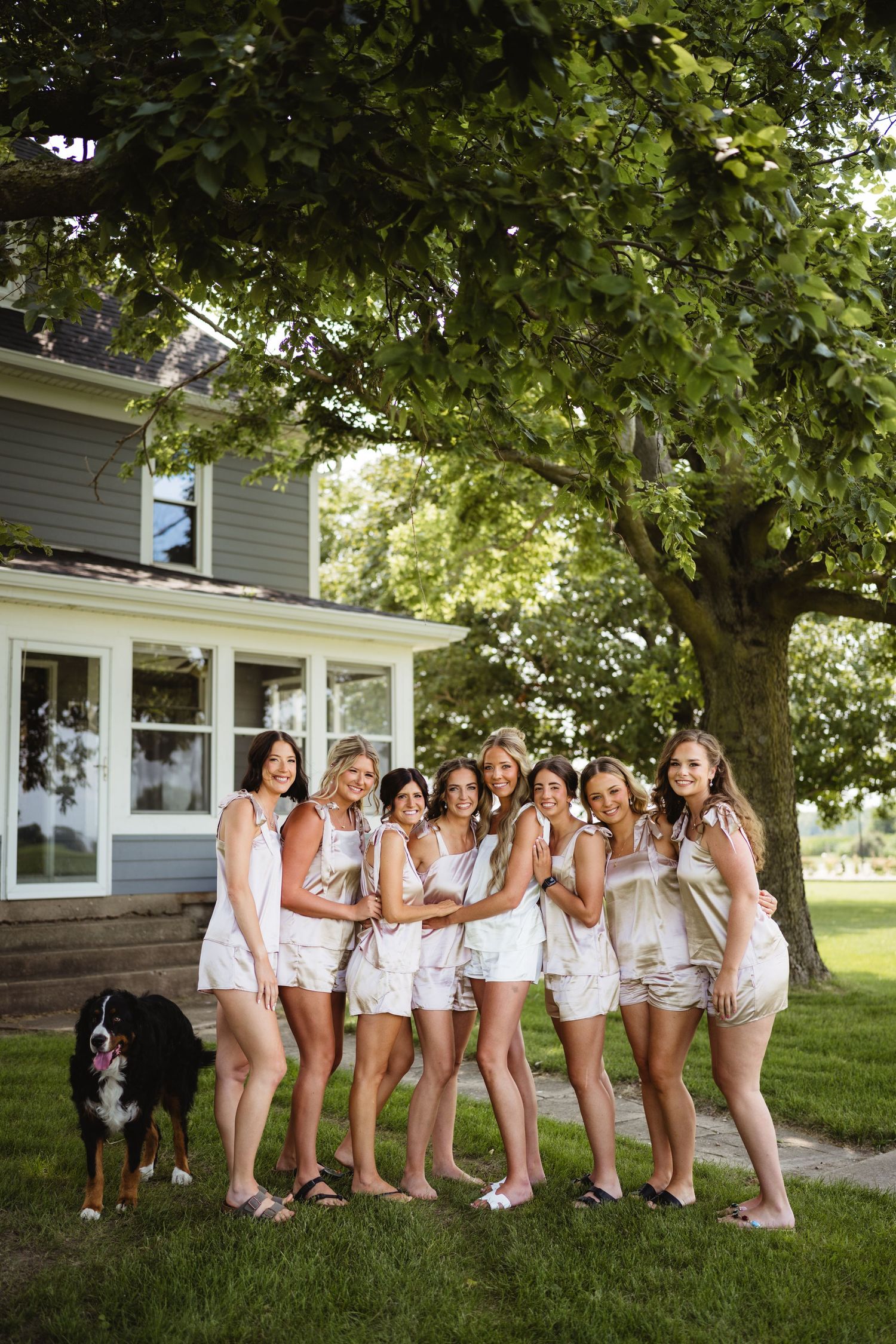 Group of women in matching white outfits posing outdoors near a tree with a Bernese Mountain Dog.