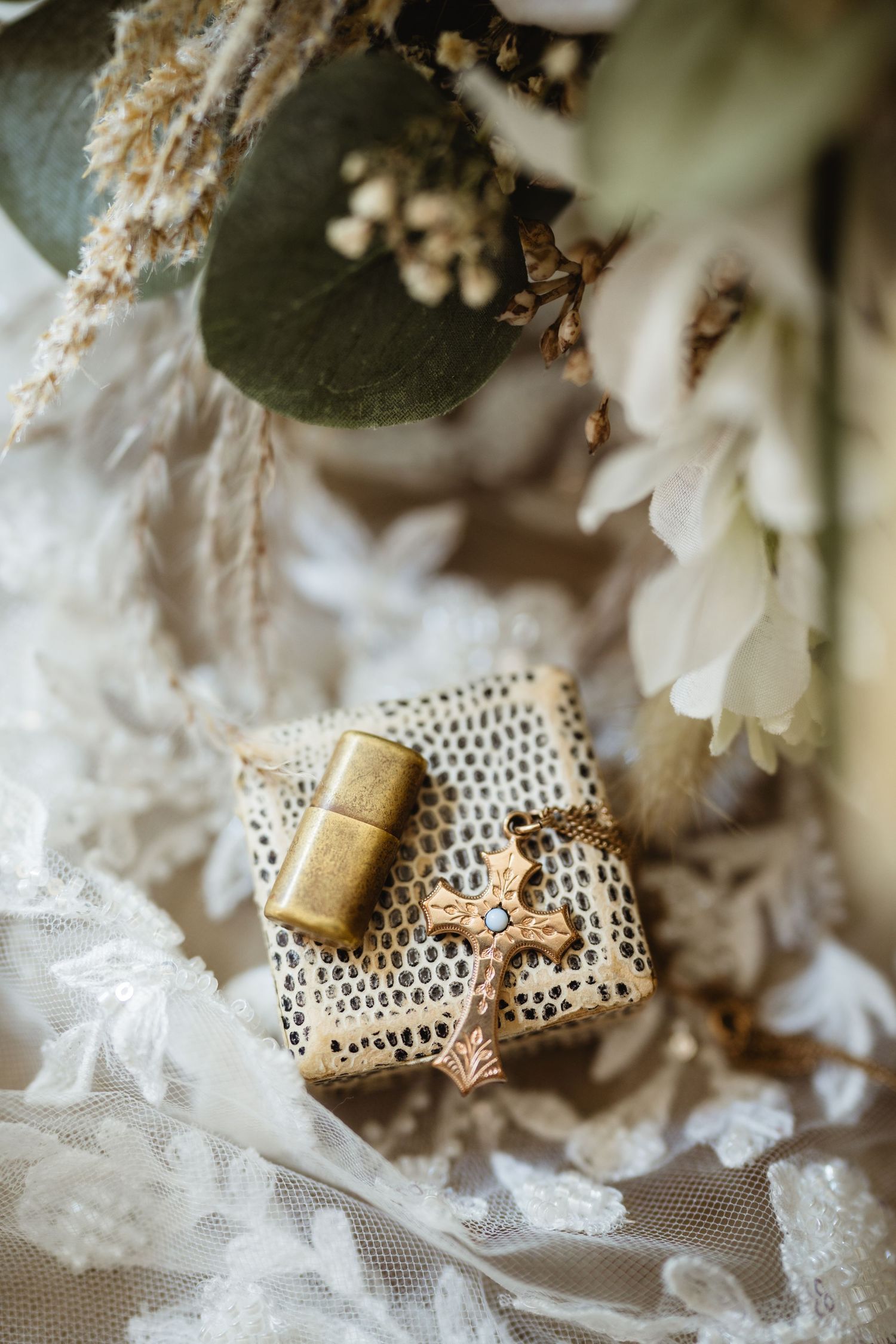 Gold cross necklace and vintage lighter on lace fabric with white flowers and eucalyptus for a wedding flat lay.