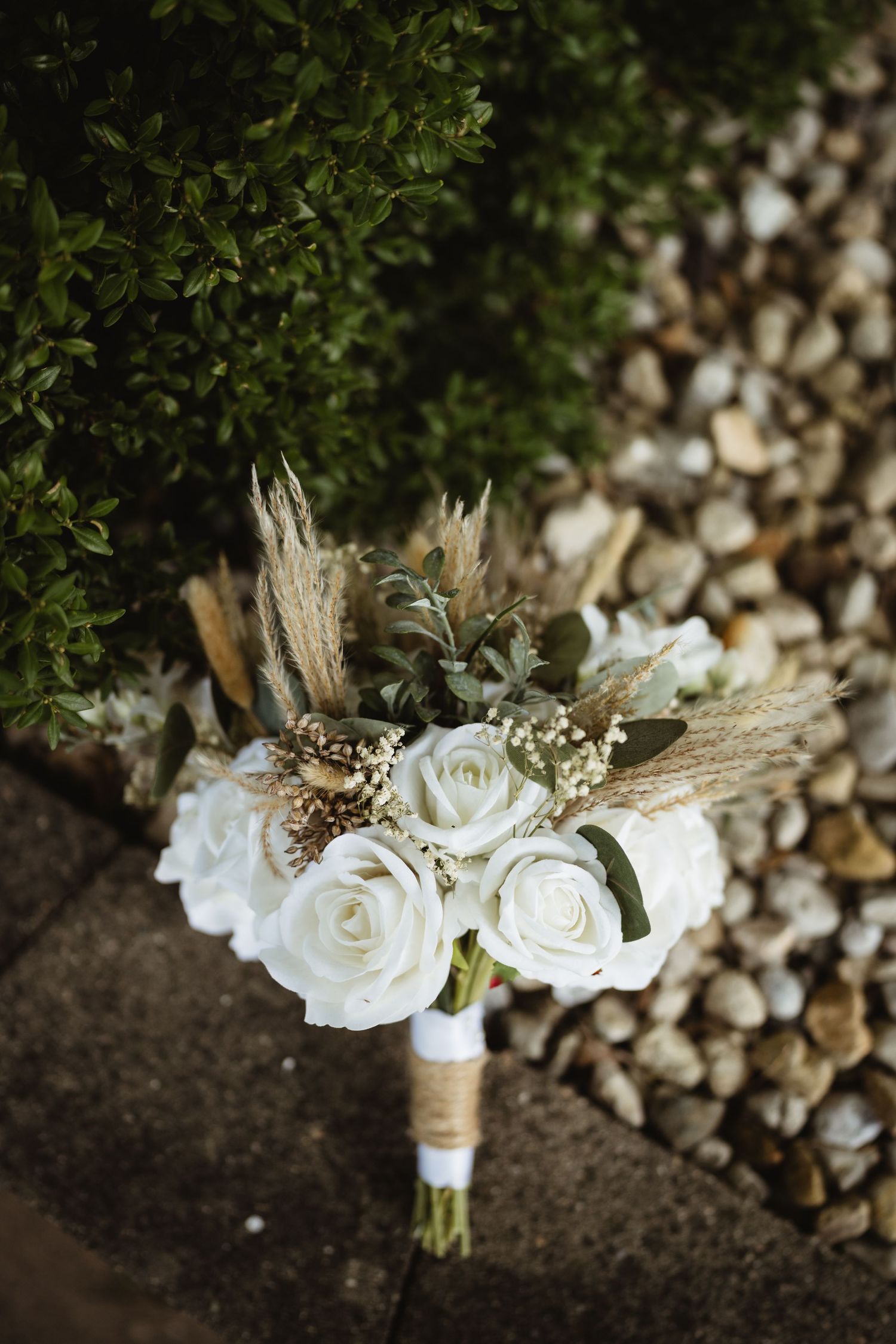 Rustic bridal bouquet with white roses, pampas grass, and eucalyptus wrapped in twine, placed on stone pavers.