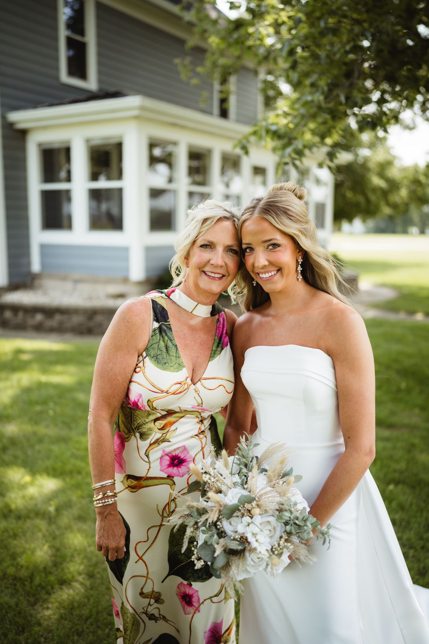 Bride in white strapless gown holding bouquet poses with woman in floral dress outside gray house.