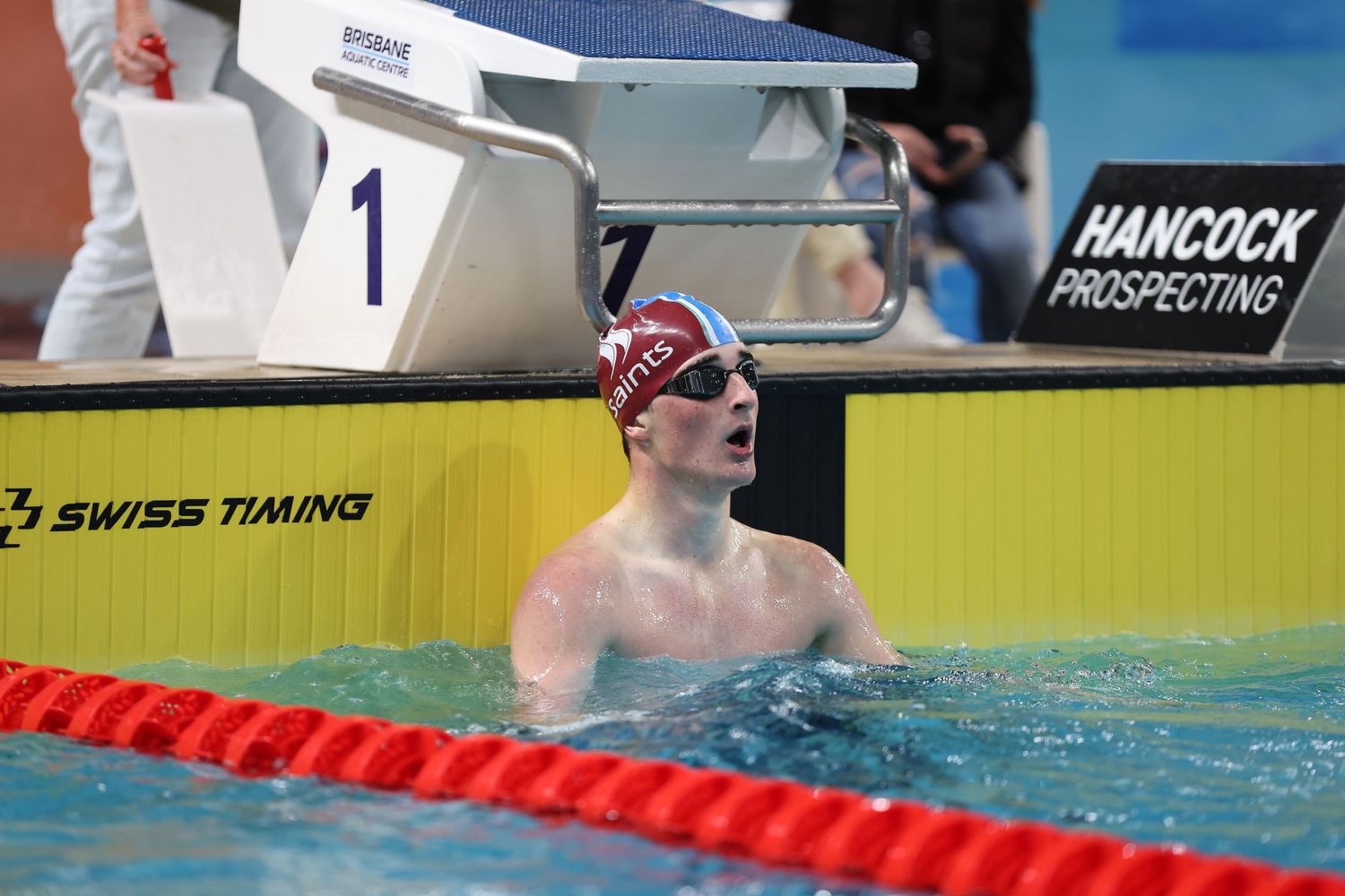 Swimmer celebrating in pool at Hancock Prospecting event, wearing red cap, with Swiss Timing scoreboard visible.