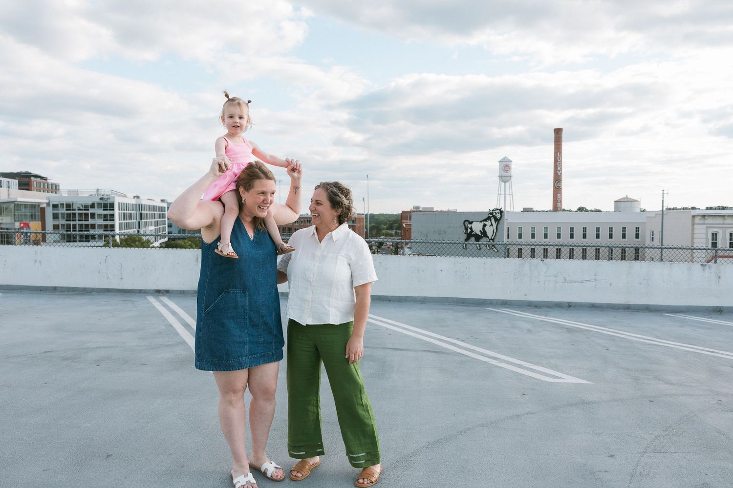 Two women and a baby girl pose on a rooftop parking lot with a city skyline backdrop.