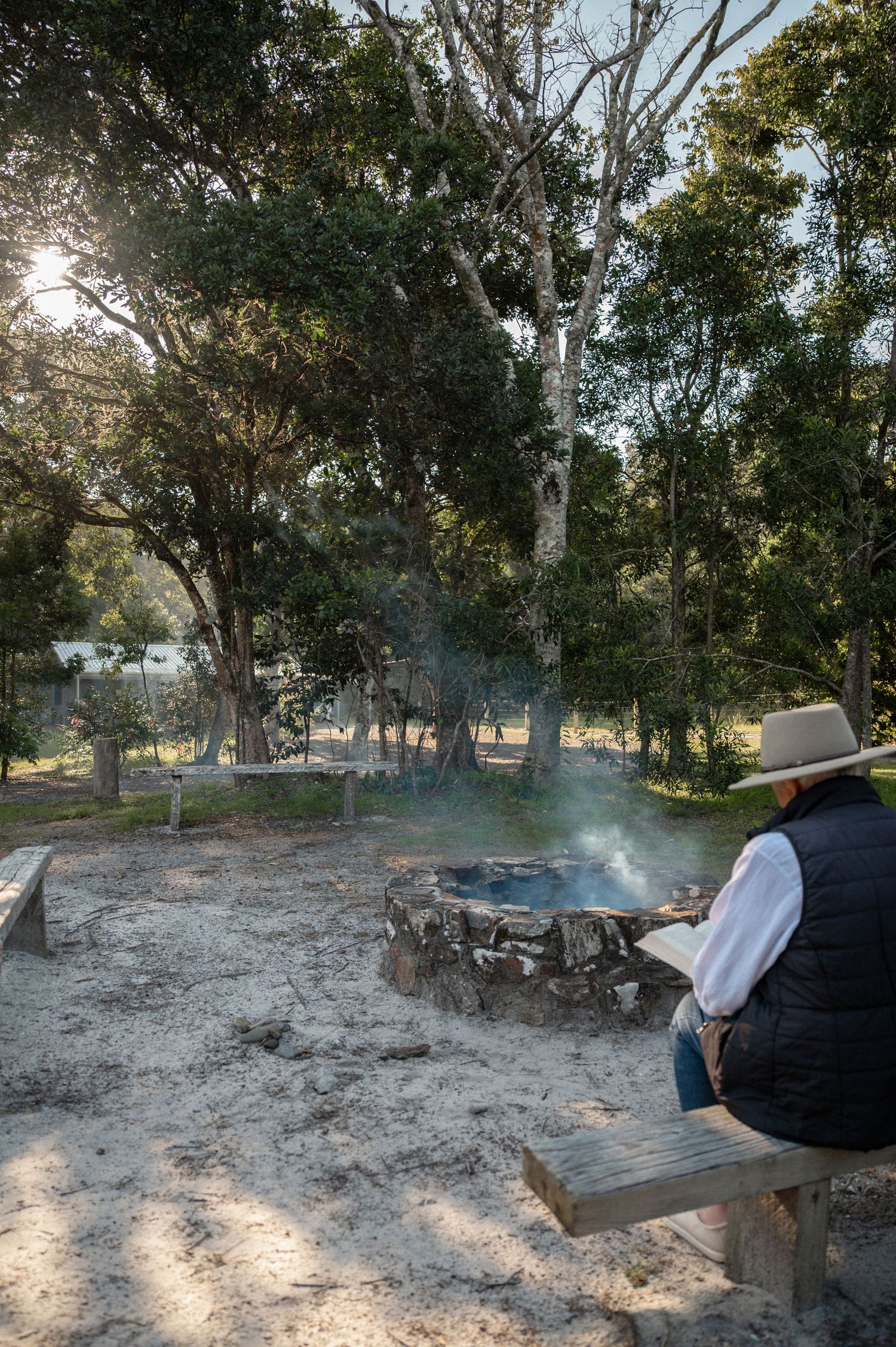 Person in hat sitting on bench by stone fire pit with smoke rising, surrounded by trees in outdoor setting.