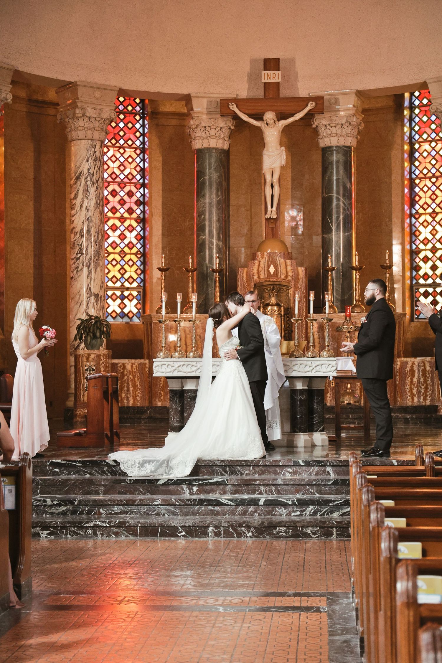 Bride and groom share first kiss at church altar during Catholic wedding ceremony with stained glass windows.