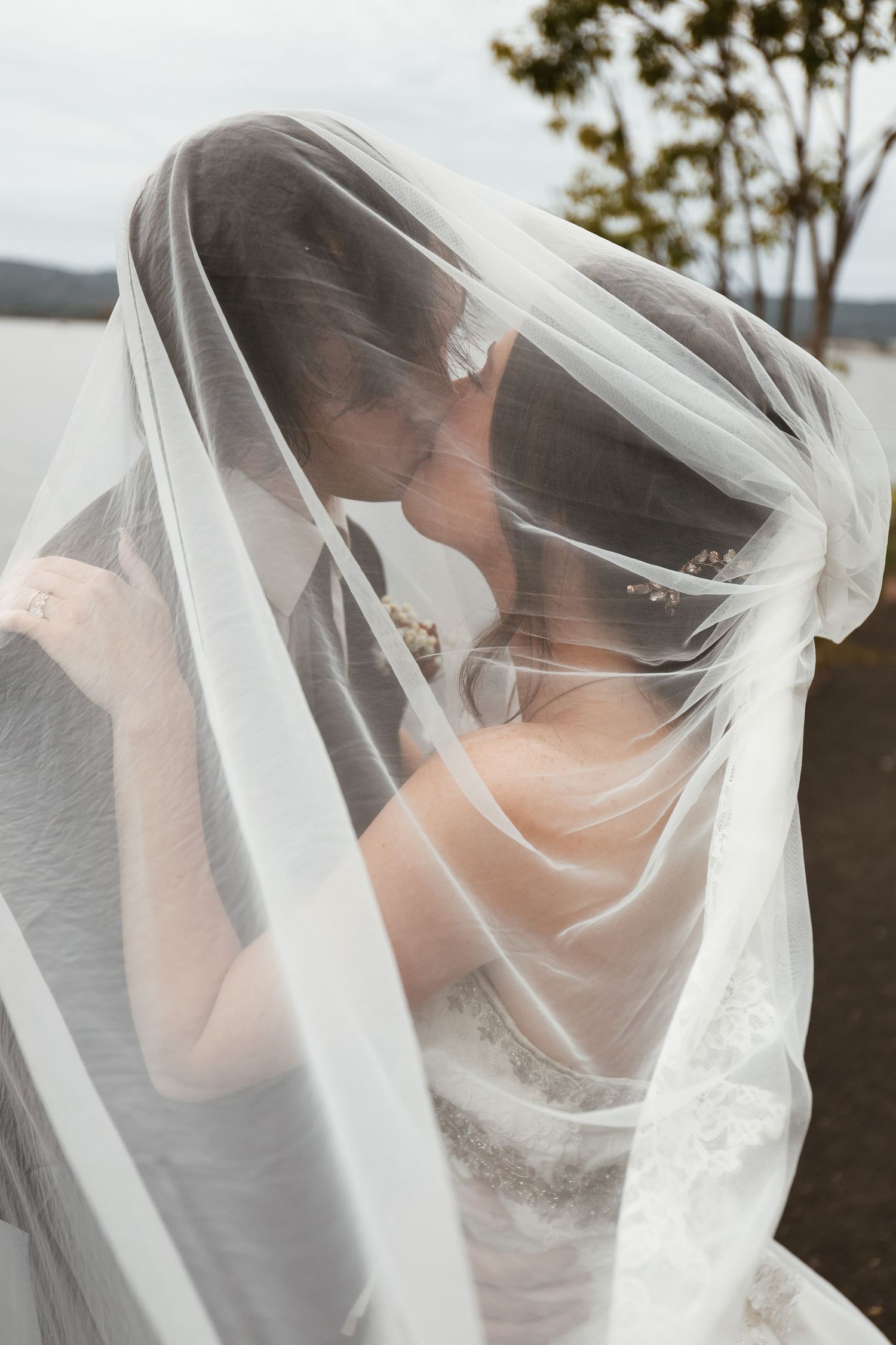 A bride and groom share a romantic kiss beneath a flowing white wedding veil outdoors near a lake.