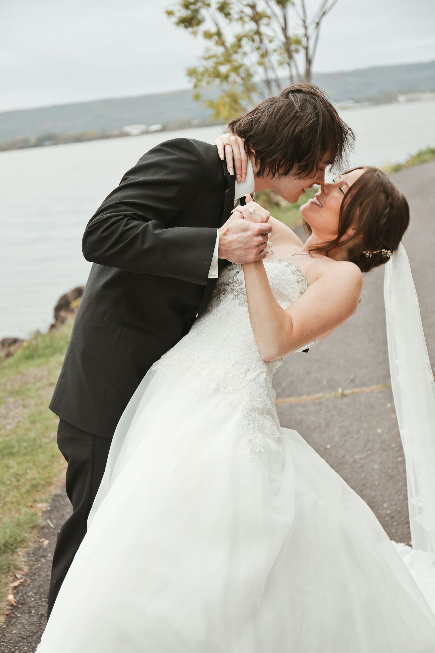 A bride and groom share a romantic kiss outdoors near a waterfront, she in a white gown, he in a black tuxedo.