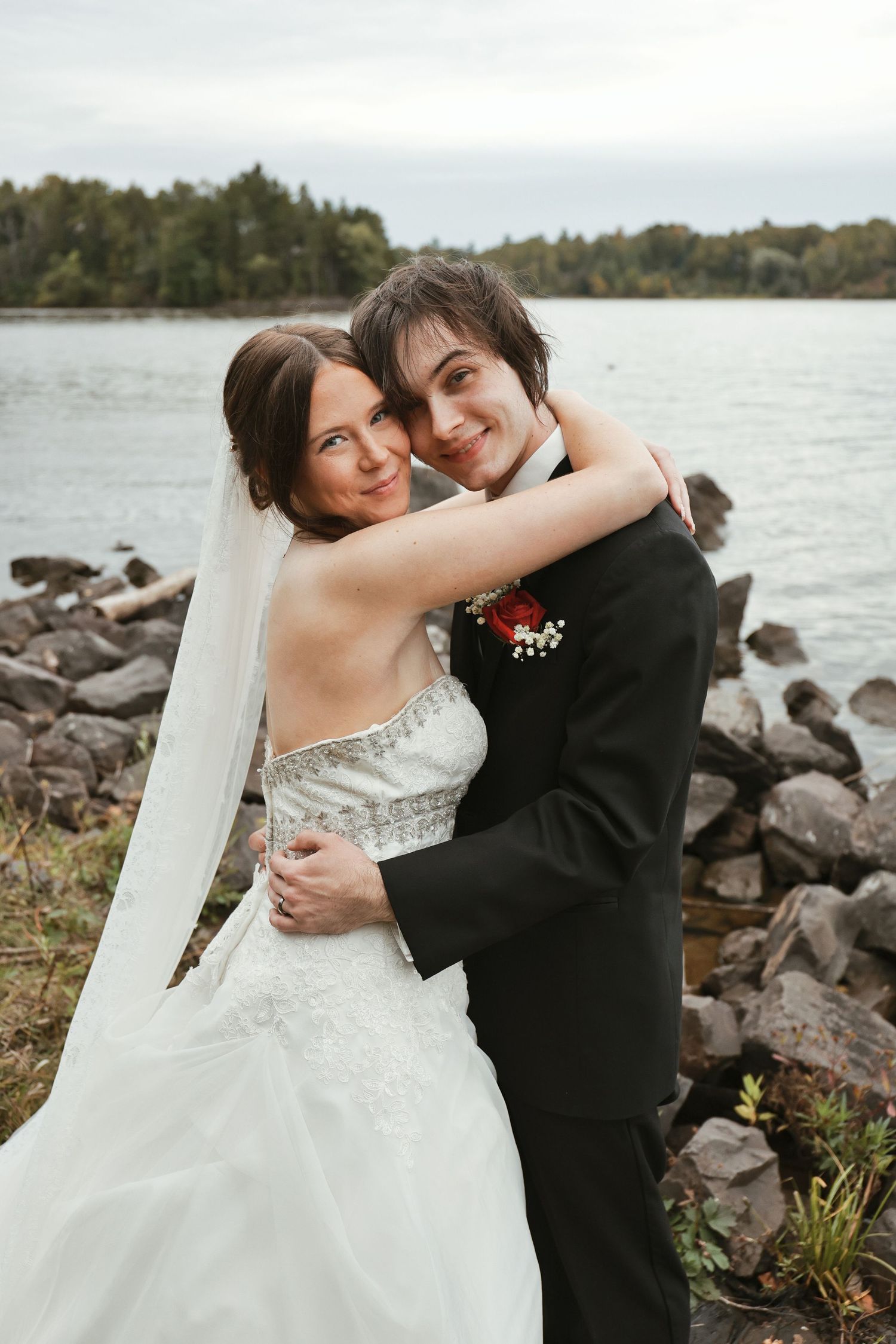 A bride and groom embrace near a rocky shoreline, smiling joyfully in wedding attire with a lake and trees in the background.