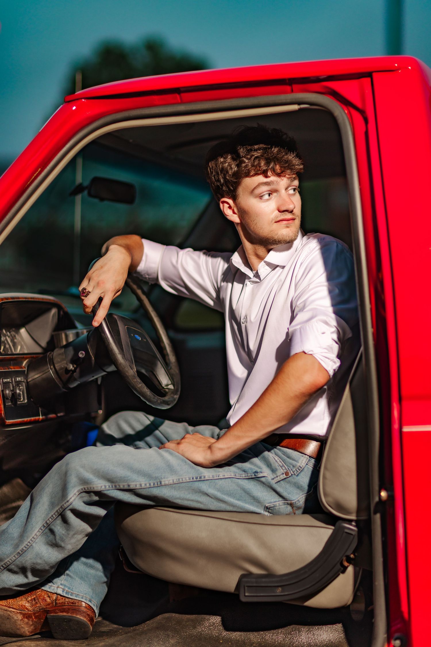 Young man in white shirt and jeans sitting in driver seat of classic red truck, looking away confidently.