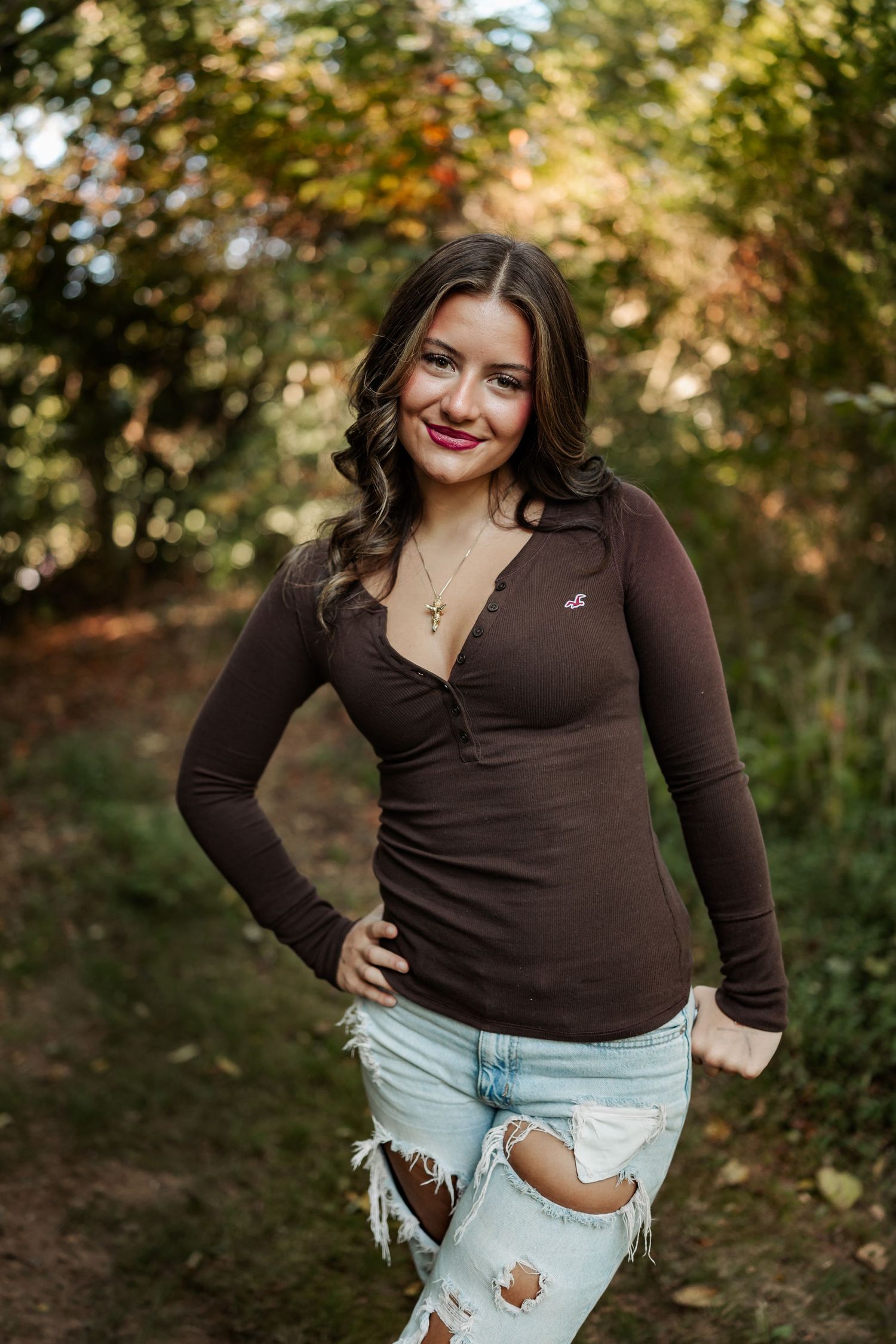 Young woman in brown long-sleeve top and ripped jeans smiling outdoors in a lush green forest setting.