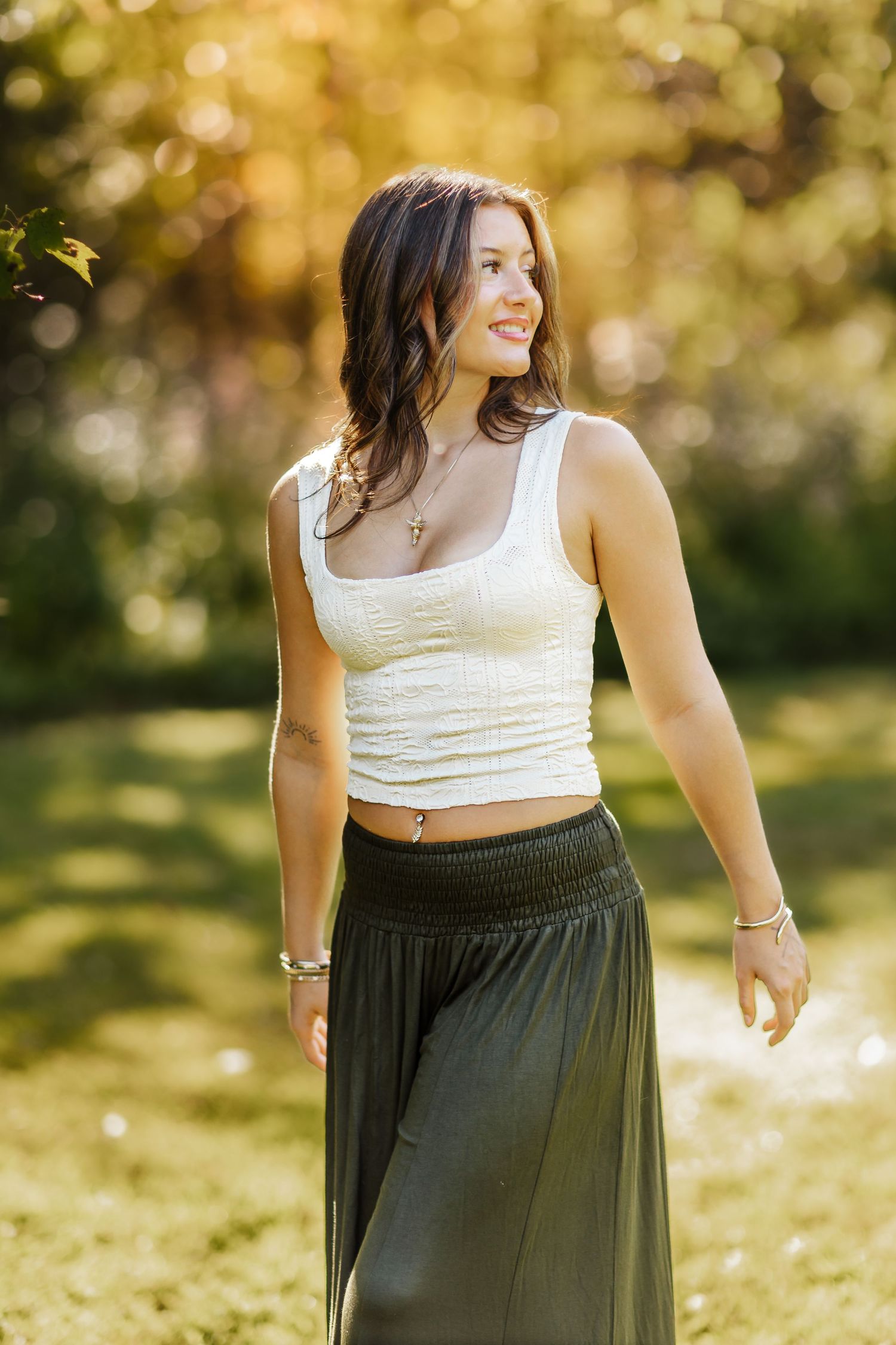 Young woman in white crop top and olive maxi skirt smiling outdoors in sunlit park setting.