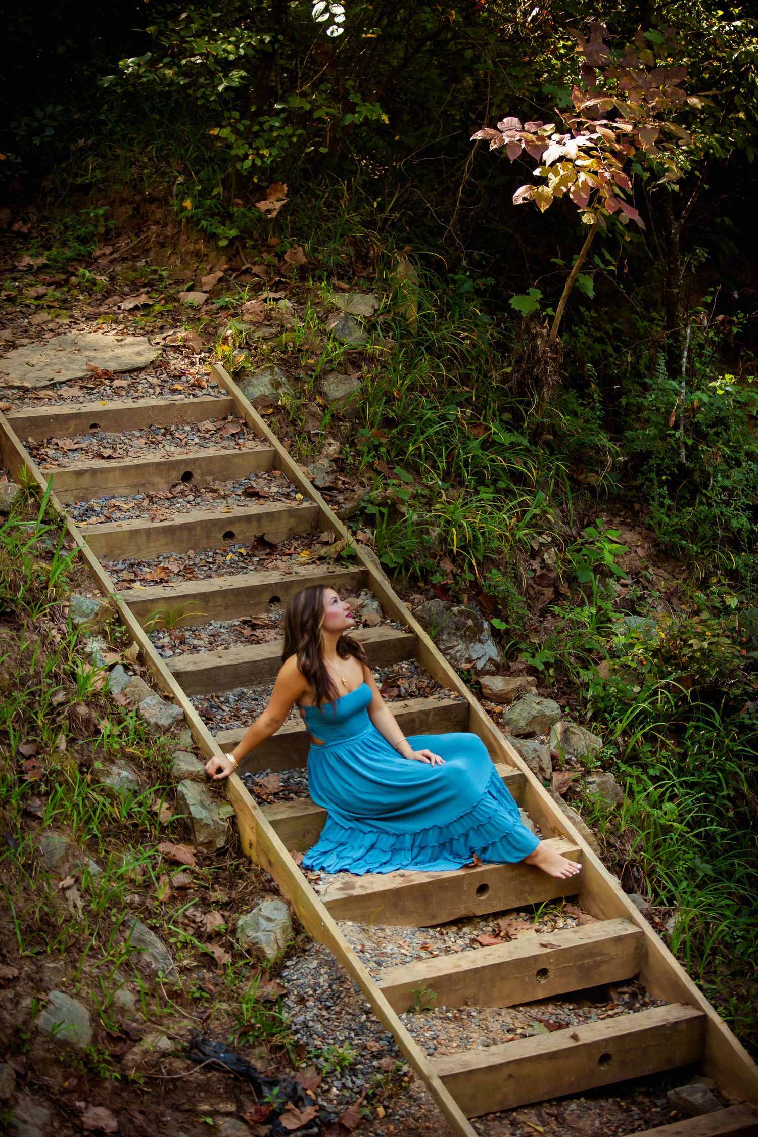 Woman in blue dress sitting on wooden outdoor stairs surrounded by lush green forest foliage.