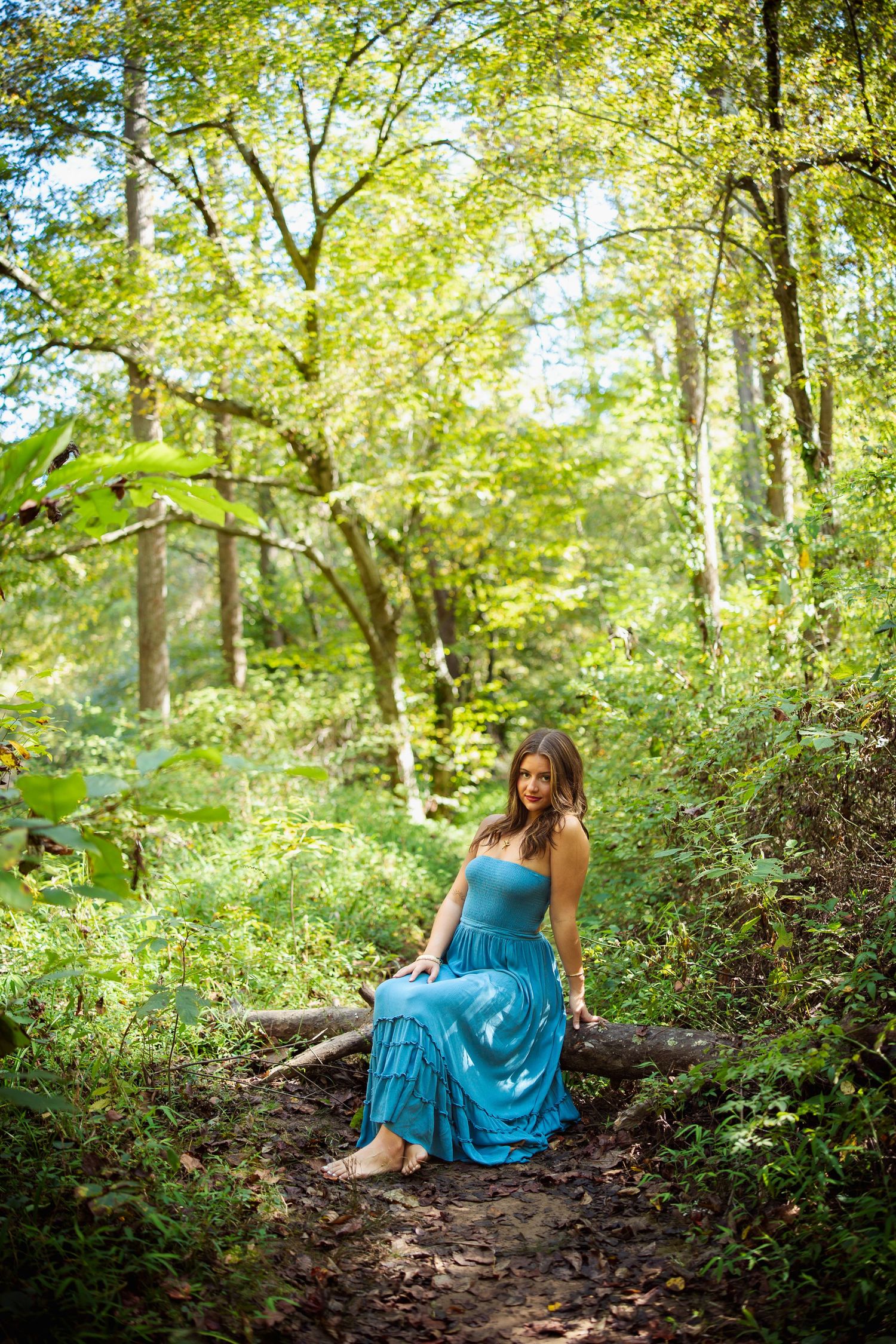 Woman in blue dress sitting on fallen log in sunlit forest.