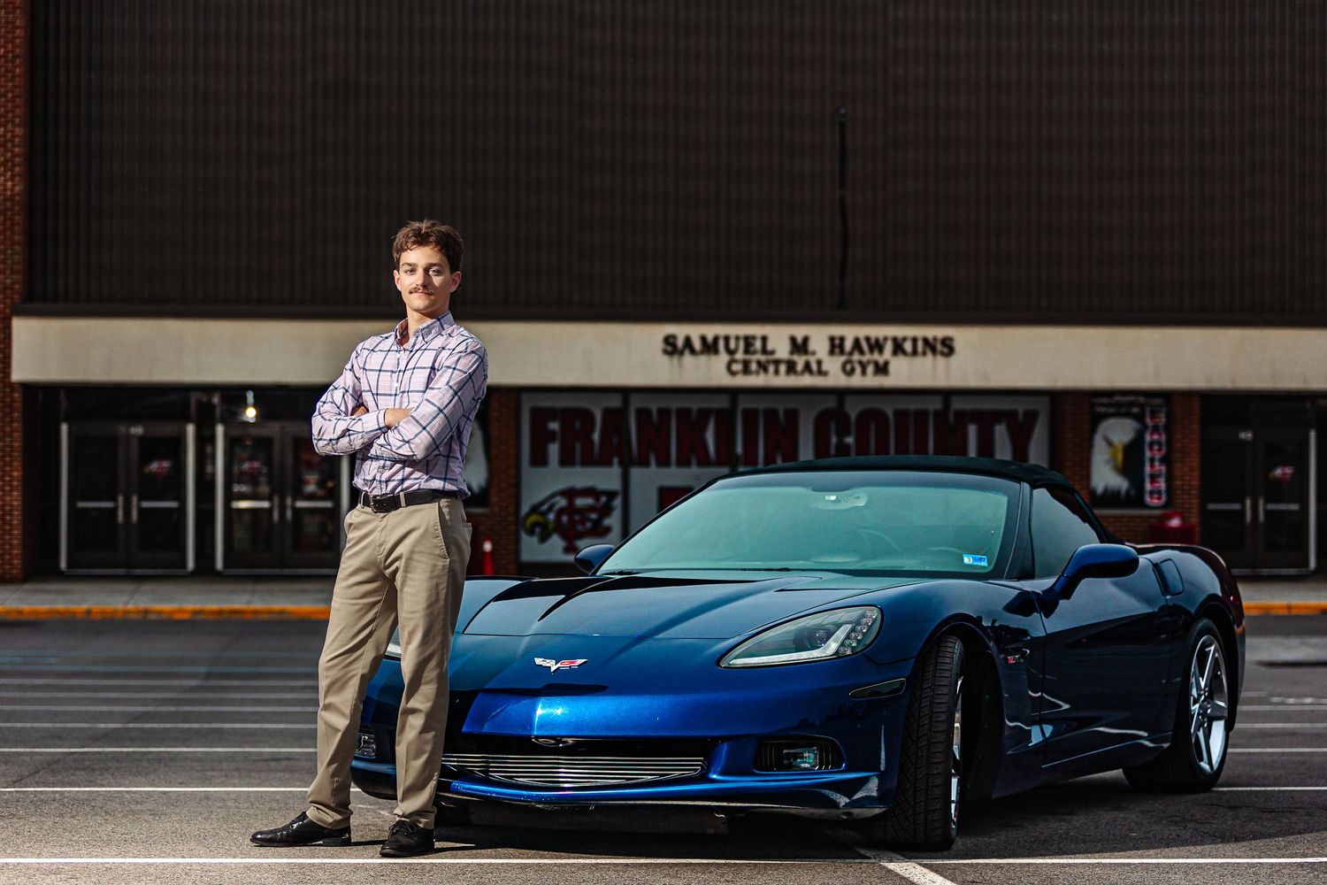 Man in dress shirt leaning against blue Corvette outside Franklin County Central Gym building.