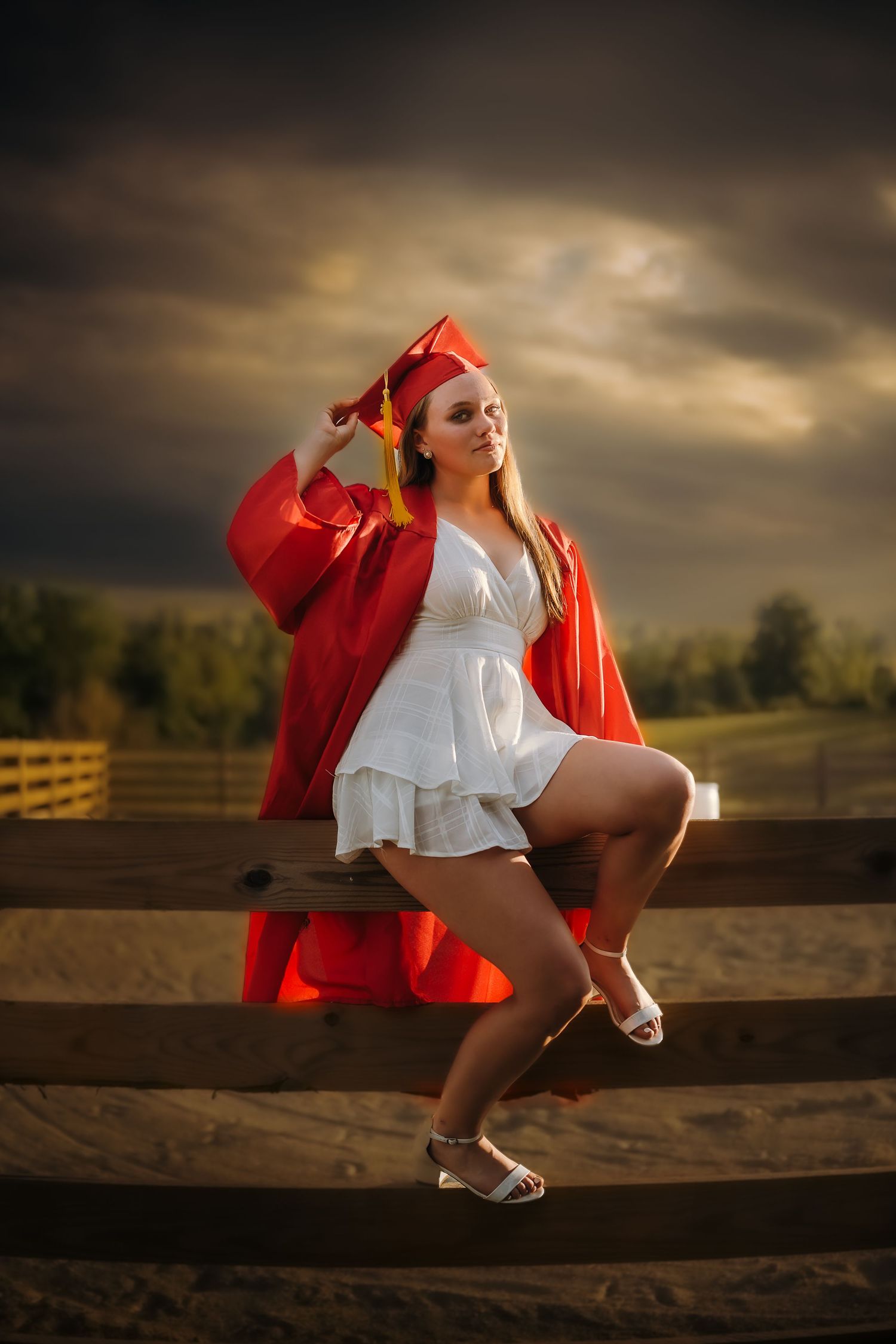 Young woman in red graduation cap and gown poses on wooden fence at sunset with dramatic cloudy sky.