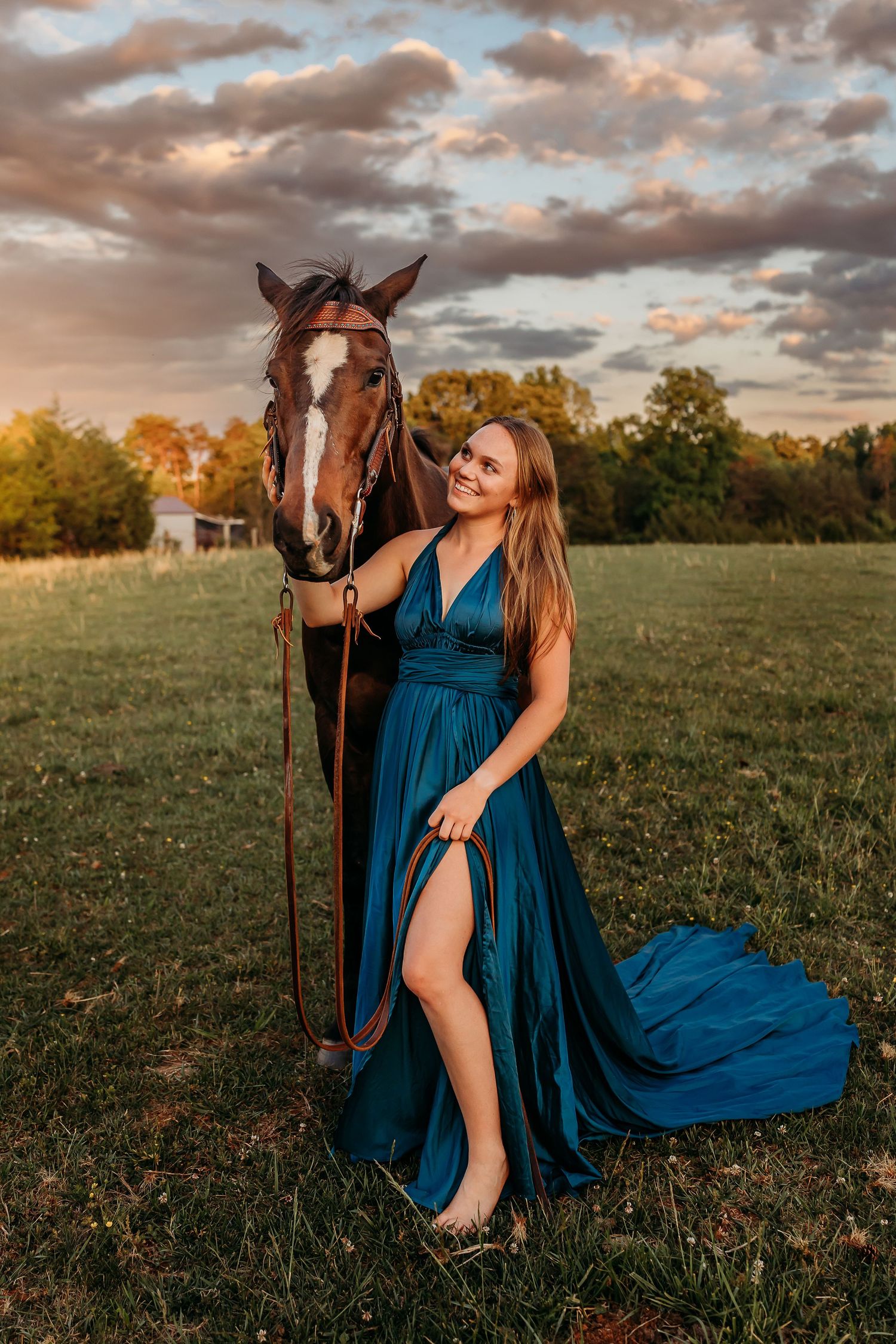 Woman in teal gown poses with brown horse at golden hour in green field.