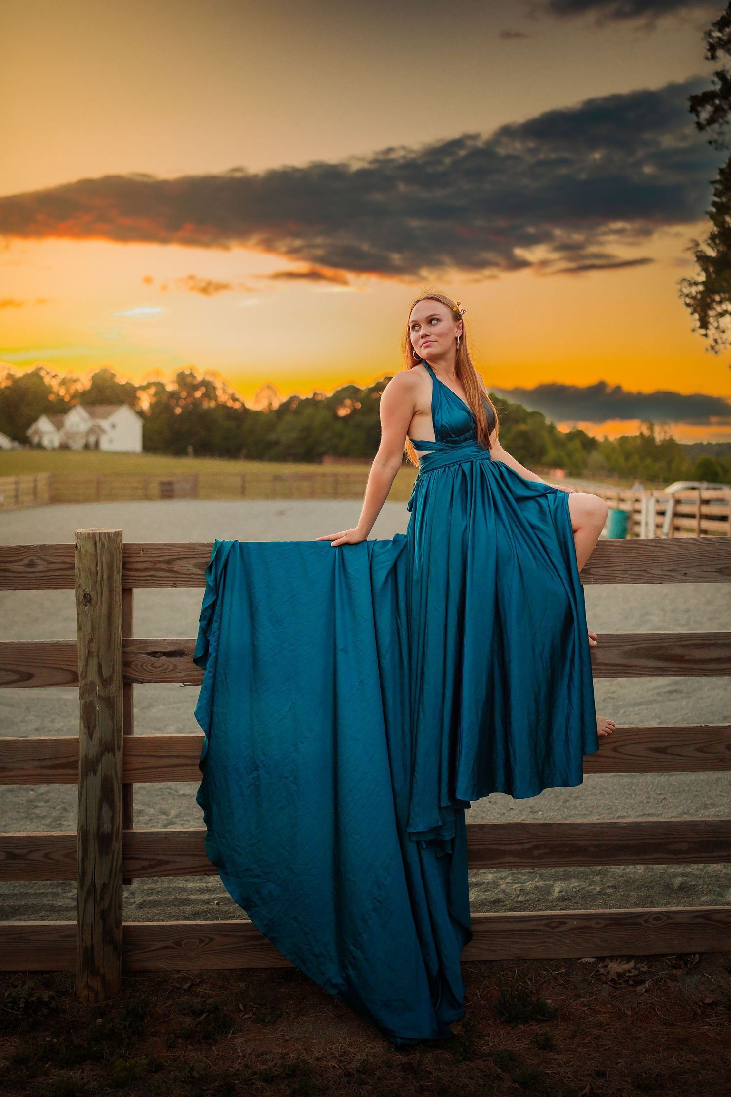 Woman in teal gown poses on wooden fence at golden sunset with dramatic clouds and rural landscape.