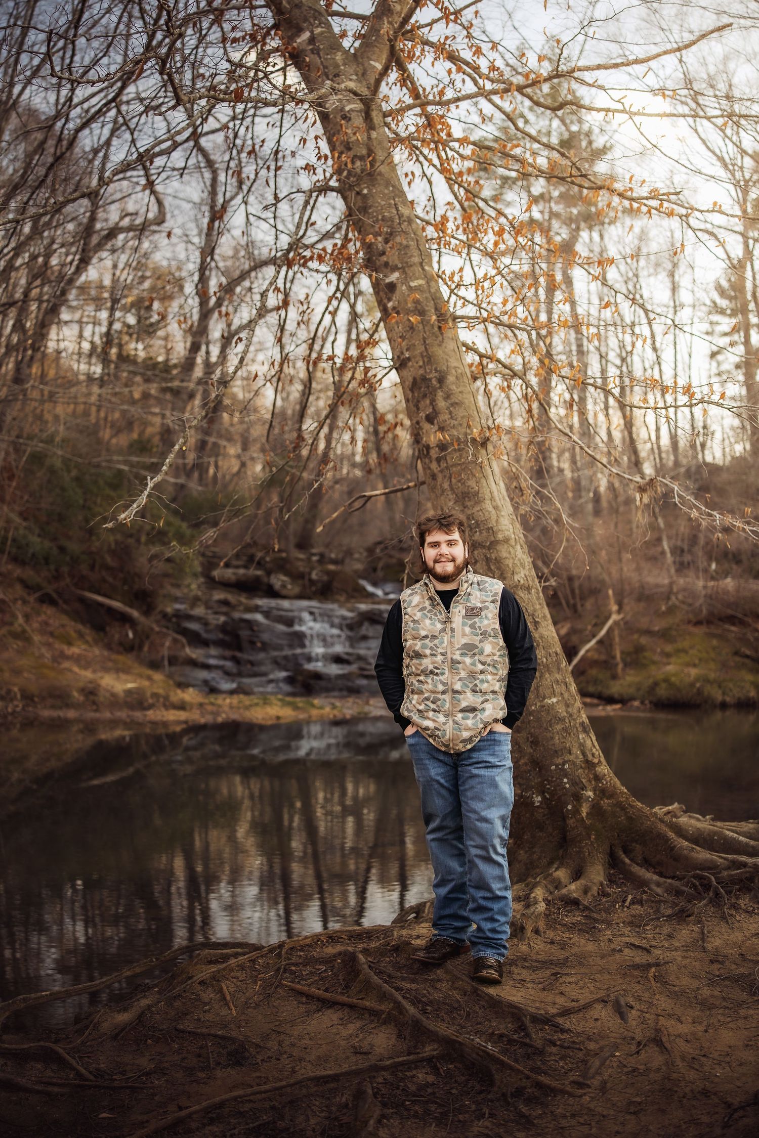 Young man in camo vest and jeans leaning against a tree by a serene woodland stream with a small waterfall.