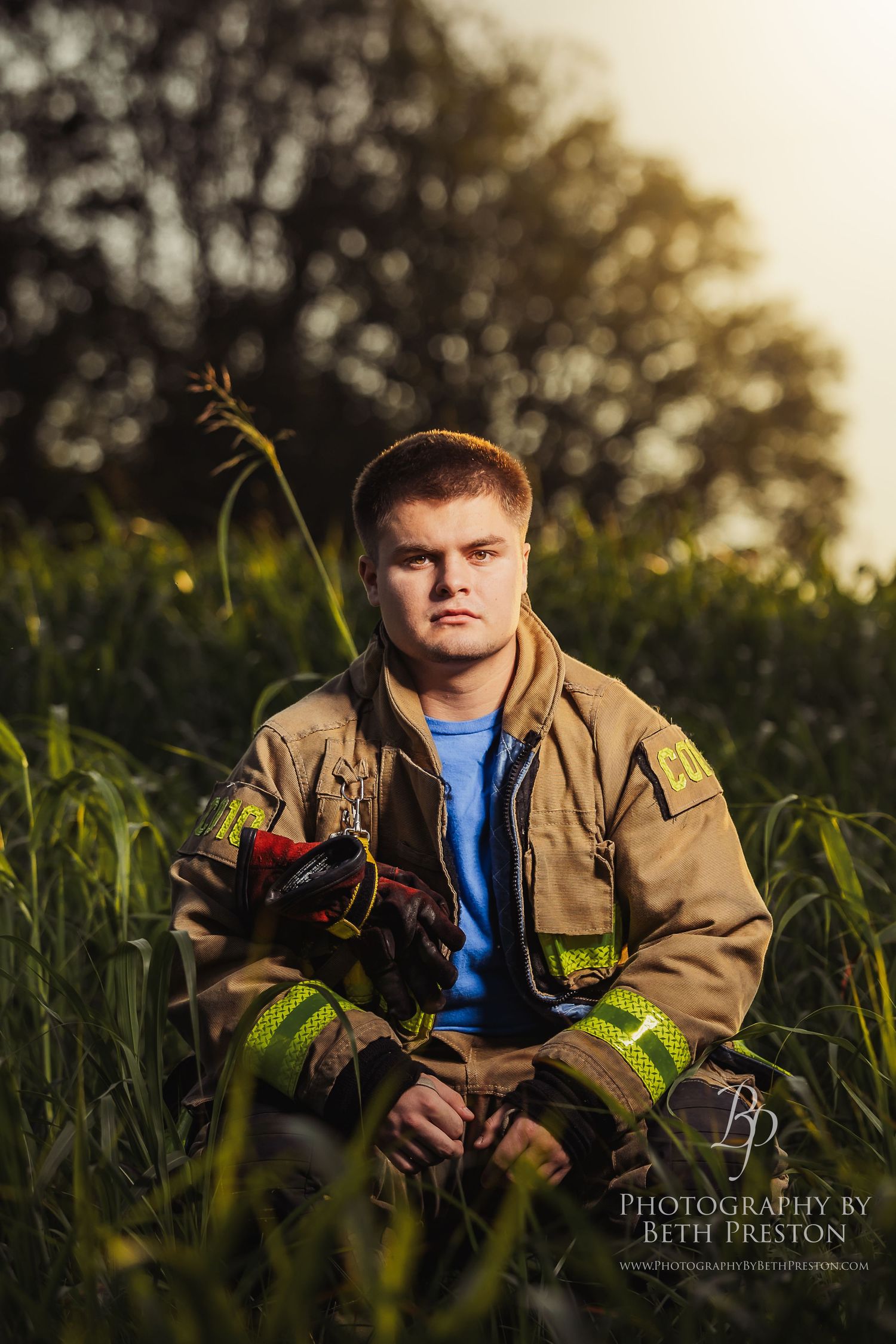 Young firefighter in turnout gear sits in a lush green field, holding gloves, in a dramatic portrait photo.