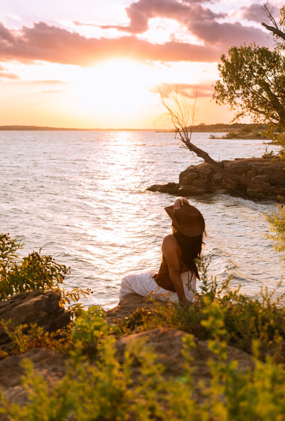 Woman in hat sitting on rocky shore watching golden sunset over calm water with dramatic cloudy sky.