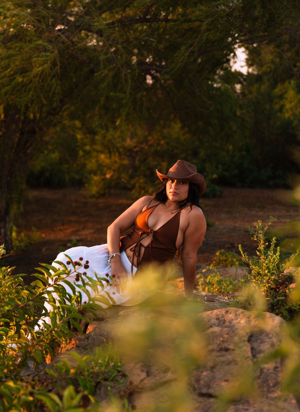 Woman in brown cowboy hat and orange top poses on rocks surrounded by lush green trees in golden hour light.