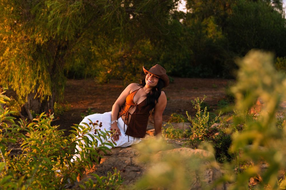 Woman in cowboy hat and brown dress sitting gracefully among lush green plants in warm golden sunset light.