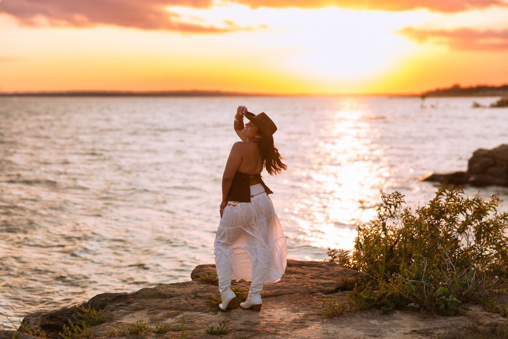 Woman in white pants and hat poses on rocky shore at golden sunset by the water.