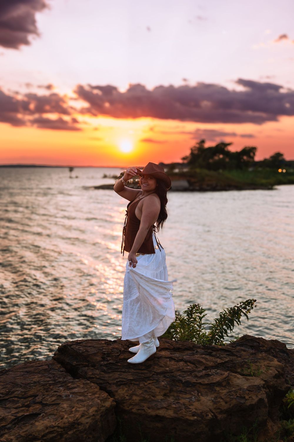 Woman in white skirt and hat stands on rocks by water during a vibrant orange and pink sunset.