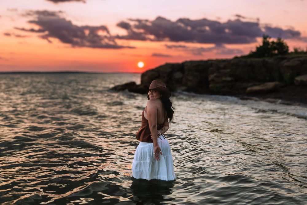 Woman in white dress standing in ocean water during vibrant sunset with rocky shore and colorful sky.