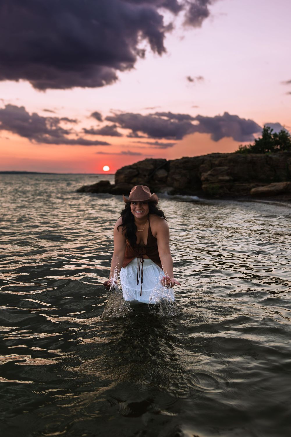 Woman in cowboy hat wading in ocean water at sunset with dramatic cloudy sky and rocky shore.