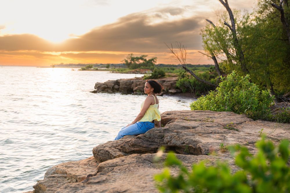 Woman sitting on rocky shoreline at golden sunset, surrounded by lush greenery and calm ocean waters.