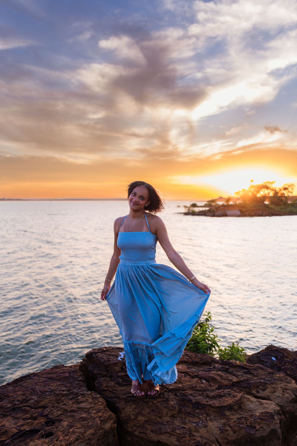 Woman in blue dress standing on rocks at sunset by the ocean.