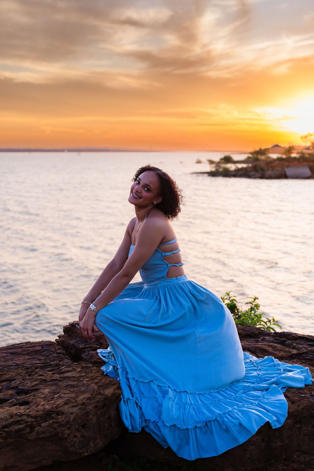 Woman in blue dress sitting on rocks by the ocean at golden sunset, smiling over her shoulder.