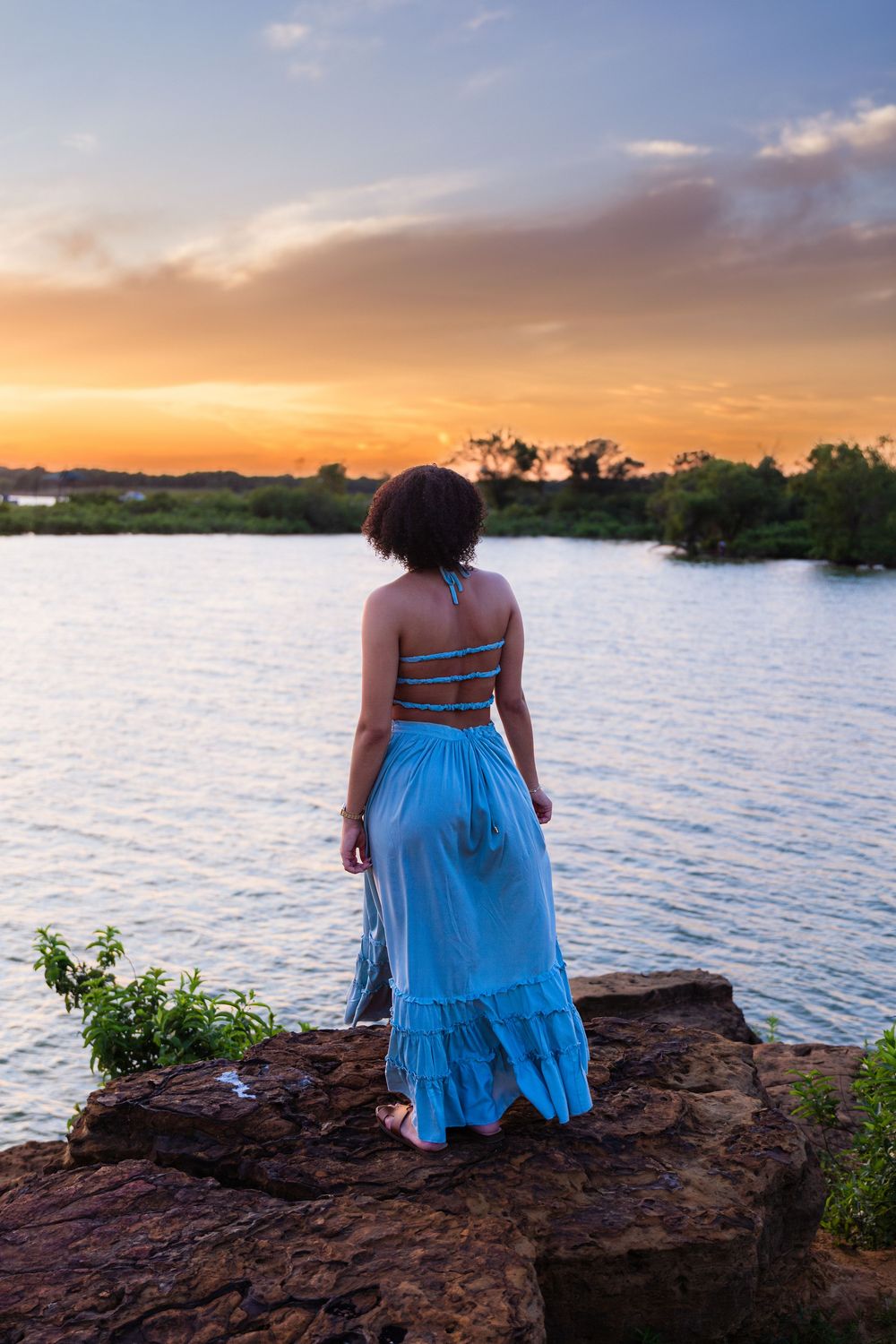 Woman in blue crop top and maxi skirt stands on rocks gazing at a vibrant sunset over a tranquil river.